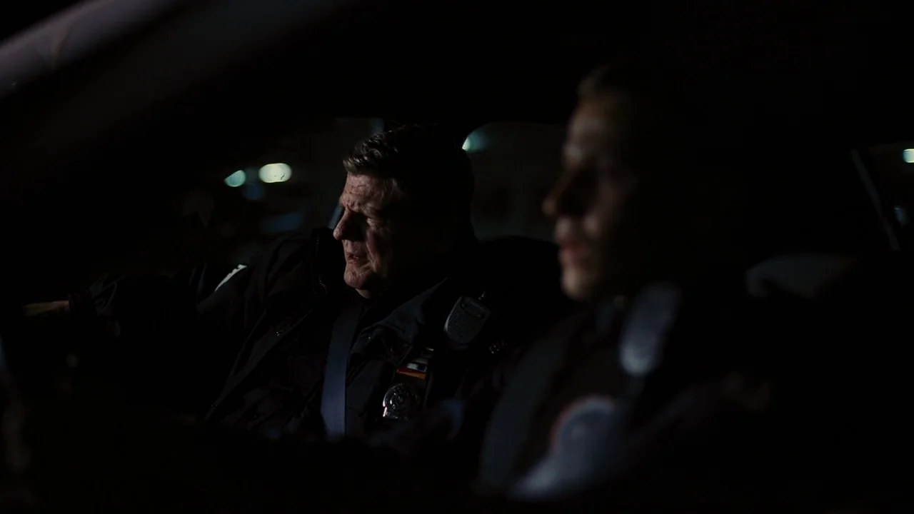 The Dark Knight Rises (2012) movie still: medium — The interior of a parked police cruiser at night is dimly lit. Two uniformed officers sit in the…