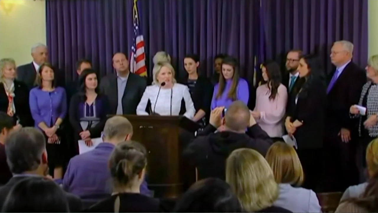 At the Heart of Gold: Inside the USA Gymnastics Scandal (2019) movie still: medium — A formal room contains a podium in front of a dark blue curtain…