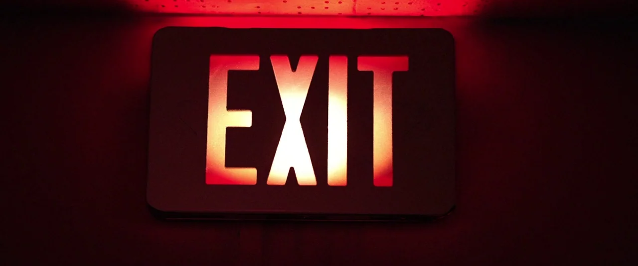 Southbound (2015) movie still: closeup — A rectangular illuminated exit sign is mounted on a dark, featureless wall surface. The bright red letters…