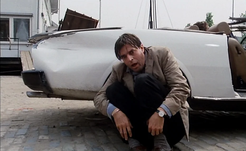 The Fourth Man (1983) movie still: medium — A paved lot sits beside a white convertible car with a damaged rear panel. A man in a tan blazer…