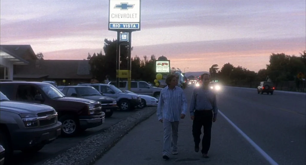 Sideways (2004) movie still: fullbody — A row of parked SUVs sits in a car lot beneath a tall Chevrolet sign. Two men walk toward the camera along…