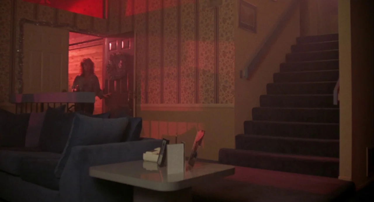 Night of the Comet movie still: wide — A dark foyer features a staircase on the right and floral wallpapered walls. A woman enters through a front…