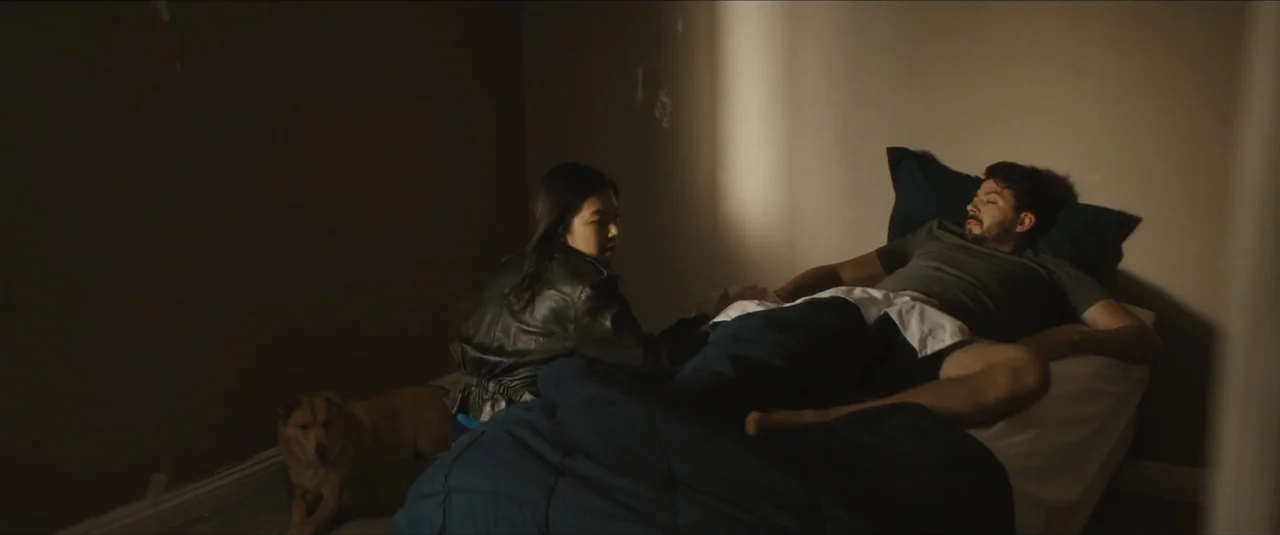 Stalker (2020) movie still: medium — A sparsely furnished bedroom with plain walls and a single bed. A woman and man rest on the bed while a dog…