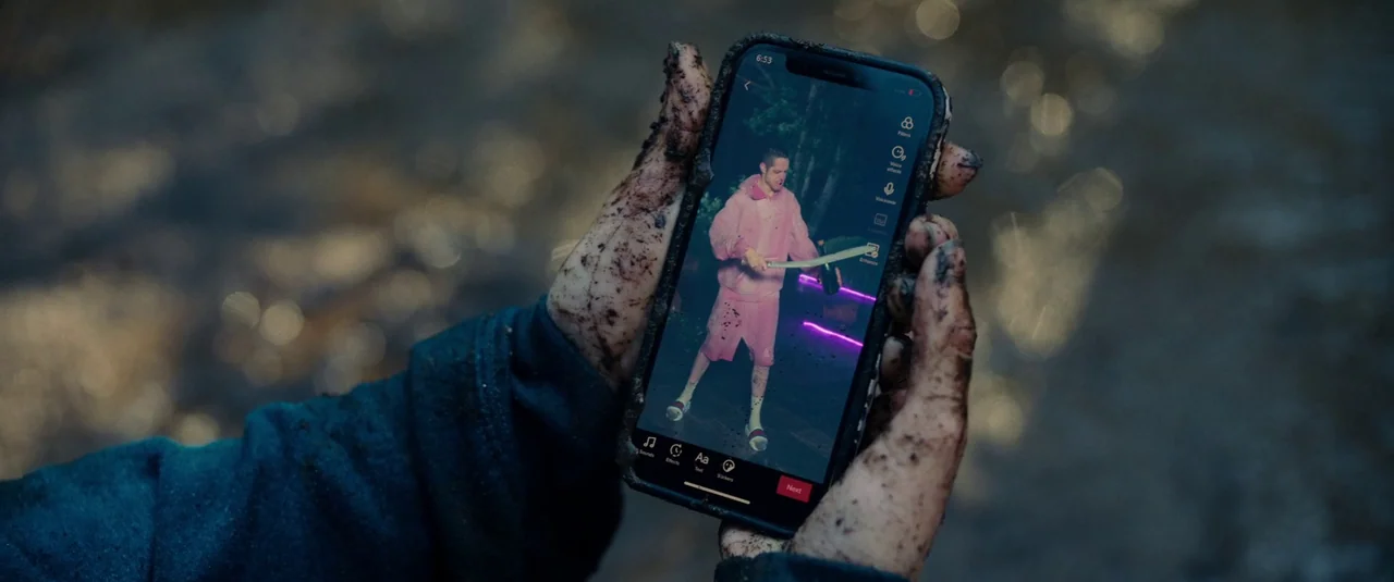 Bodies Bodies Bodies (2022) movie still: closeup — A pair of mud-caked hands holds a smartphone displaying a video in a natural environment. The…