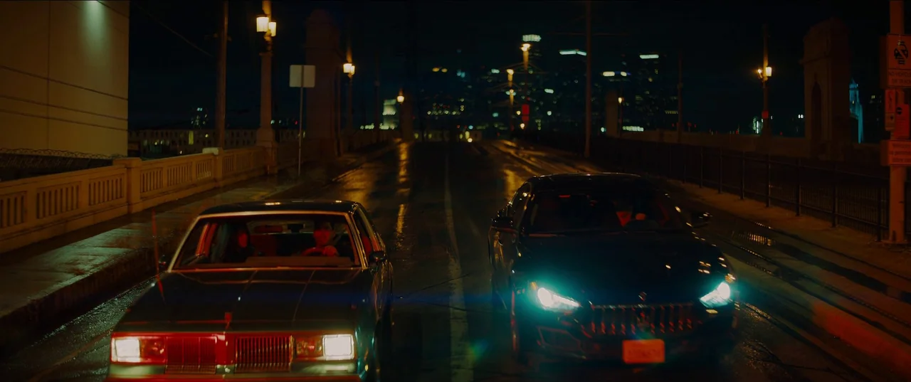 Night Teeth (2021) movie still: wide — A wet asphalt bridge at night features two cars driving in parallel lanes. City lights glow in the distance…
