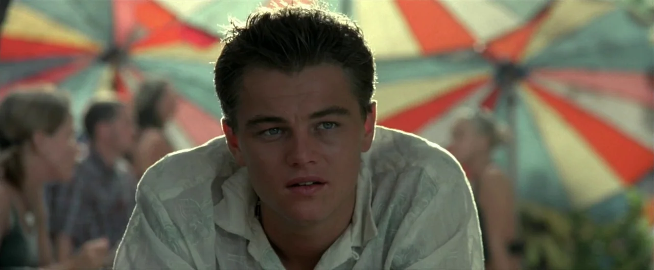 The Beach (2000) movie still: closeup — A sunny outdoor setting features large colorful parasols providing shade for people. A young man sits in the…