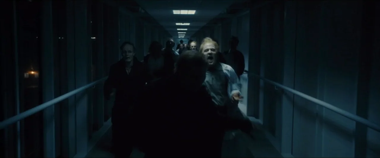 World War Z (2013) movie still: medium — A long, dimly lit indoor walkway features fluorescent ceiling lights and white metal railings. A small…