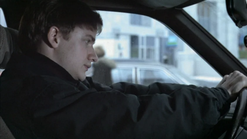 71 Fragments of a Chronology of Chance (1994) movie still: medium — A man sits in the driver seat of a vehicle during the day. He grips the steering…