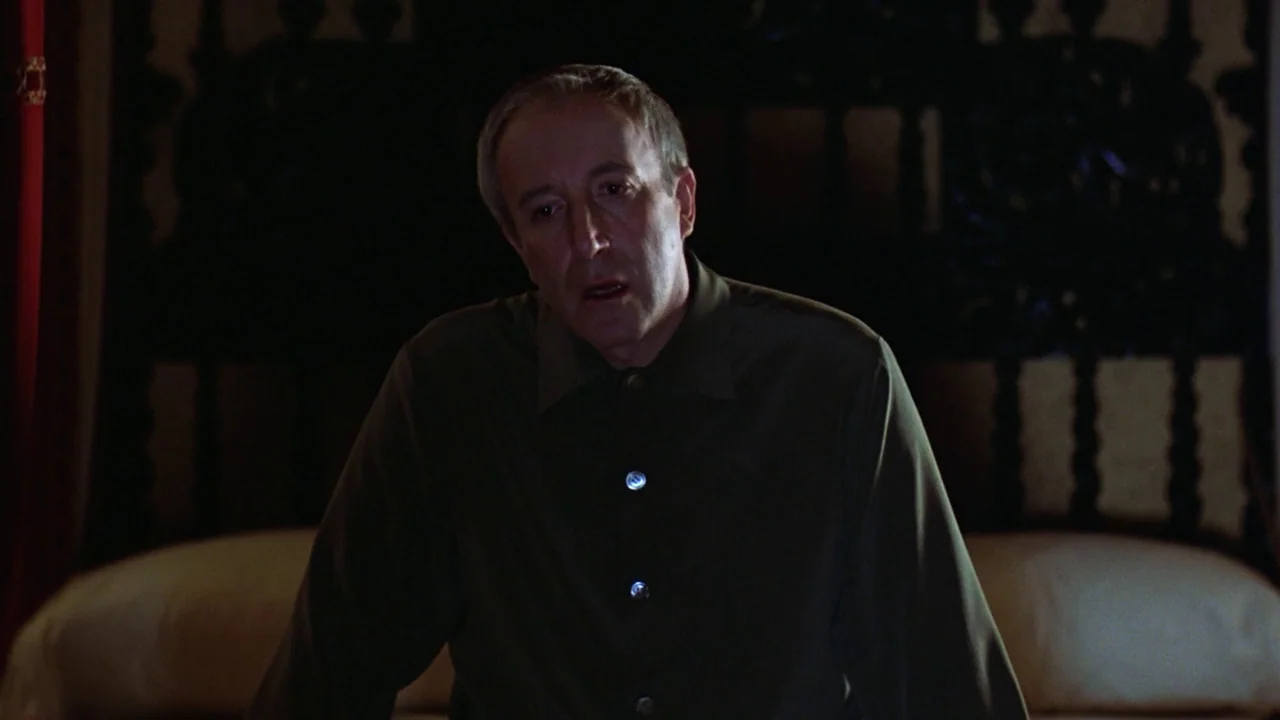 Being There (1979) movie still: medium — A dimly lit bedroom features an ornate, dark wooden headboard behind a person. The man in a green shirt…