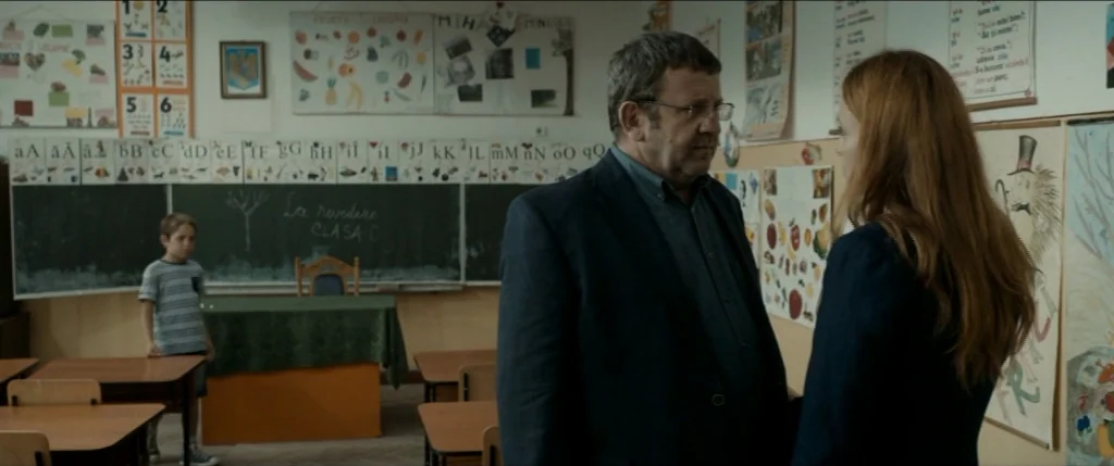 Graduation (2016) movie still: medium — A primary school classroom with educational posters, a blackboard, and wooden desks. A man and a woman face…