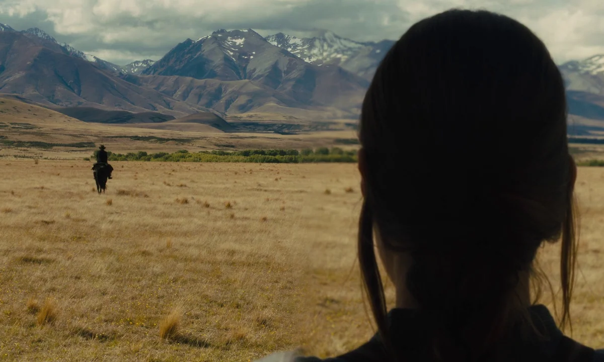 Slow West (2015) movie still: wide — A vast, dry grassy plain stretches toward snow-capped mountains under a cloudy sky. A person stands in the…