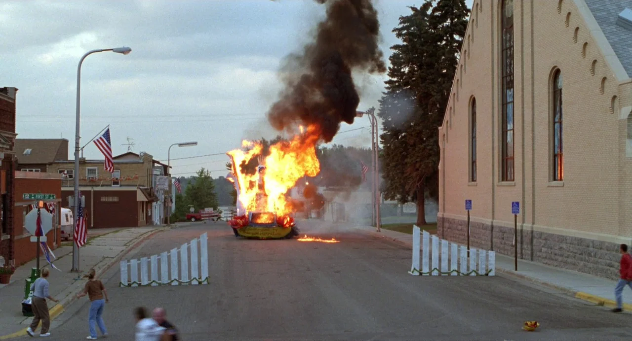 Drop Dead Gorgeous (1999) movie still: wide — A small town street features a brick church on one side and commercial storefronts opposite. A burning…