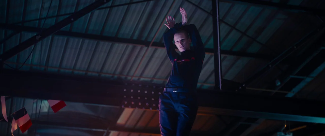Titane (2021) movie still: medium — The interior of a high-ceilinged hangar with visible steel support beams overhead. A young man stands centered…
