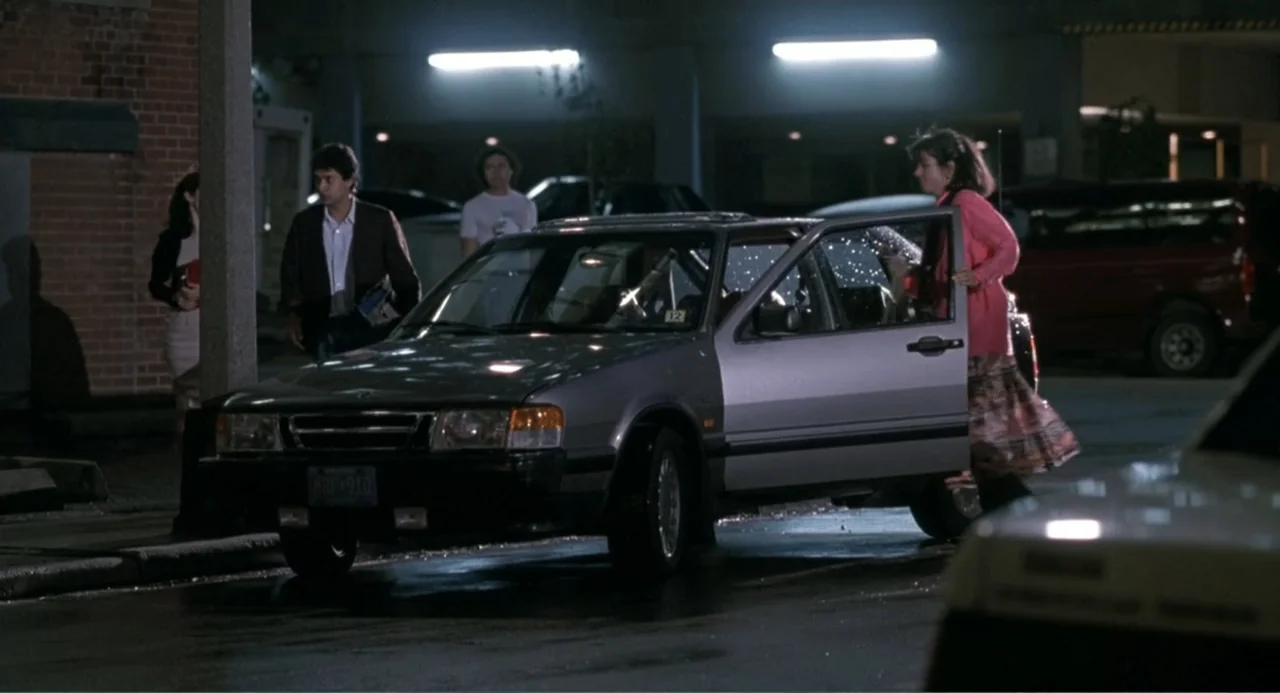 F/X2 (1991) movie still: medium — A dark urban parking lot at night with overhead fluorescent lights illuminating the pavement. A woman steps out of…