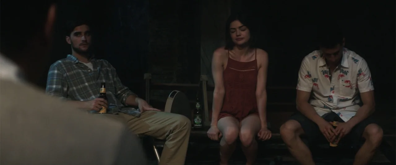 Truth or Dare (2018) movie still: medium — Three young adults sit on a wooden bench inside a dark room at night. They each hold a beer bottle while…