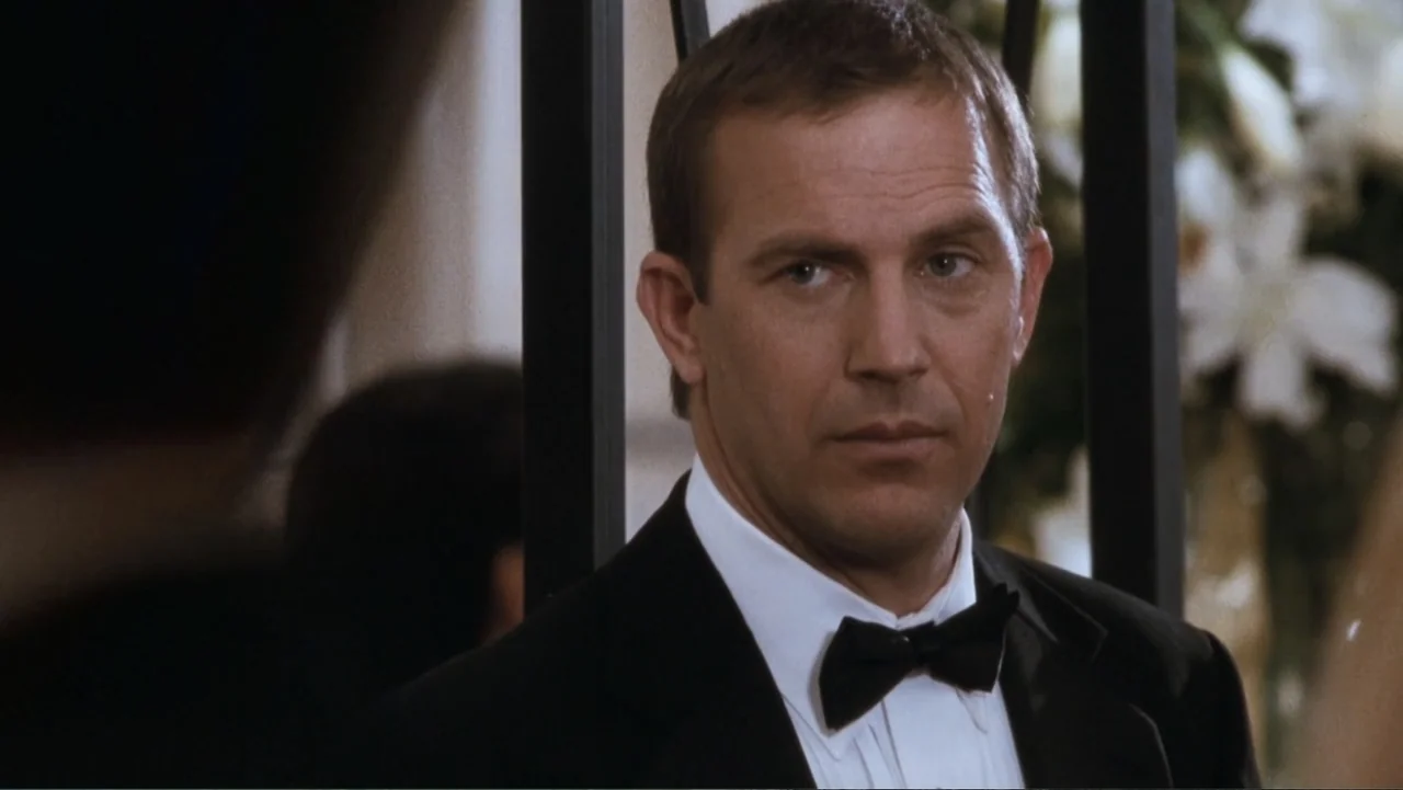 The Bodyguard (1992) movie still: medium — An interior space features black vertical railings and white floral arrangements in the background. A man…