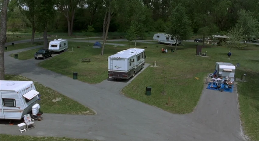 About Schmidt movie still: establishing — A rural RV campground features several parked campers and trailers on gravel paths. Trees surround the…