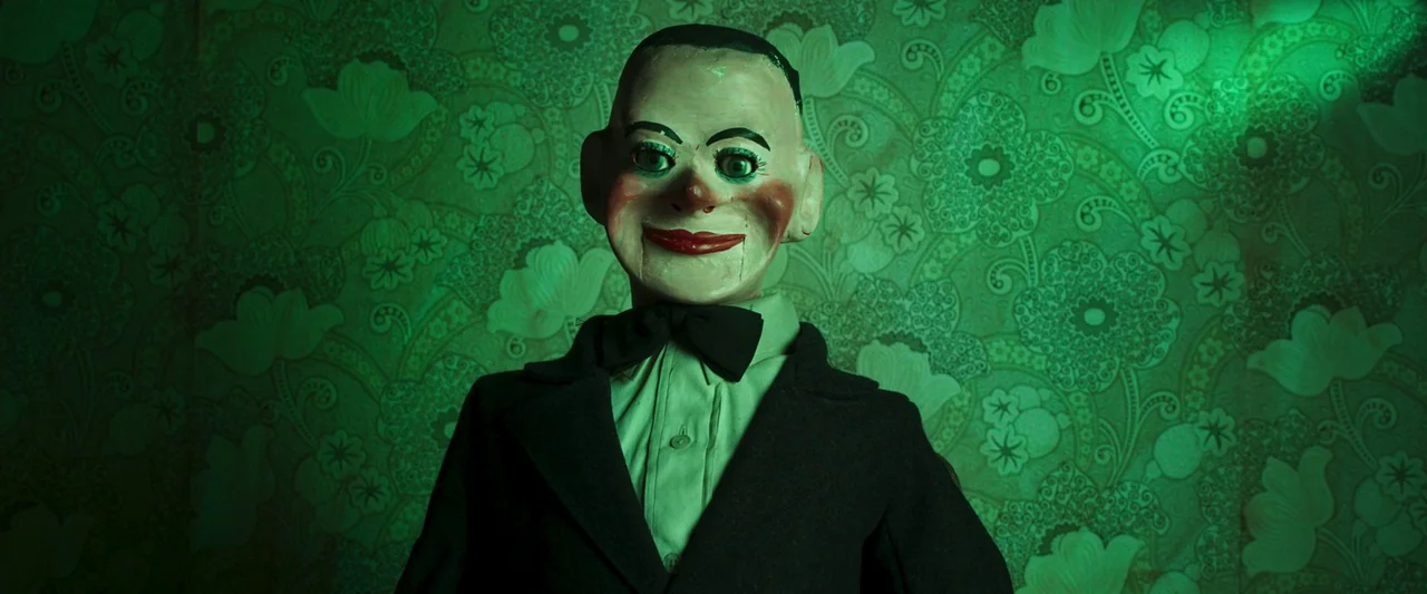 Stopmotion (2024) movie still: medium — A vintage ventriloquist dummy stands against green floral patterned wallpaper in a dark room. The dummy…