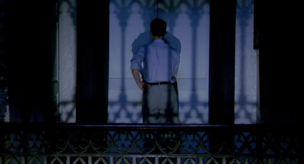 In a Glass Cage (1986) movie still: fullbody — A man stands on a balcony behind a patterned iron railing at night. The railing casts long, sharp…