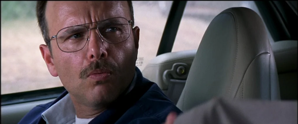 Memento (2000) movie still: closeup — A man in wire-rimmed glasses sits in the driver seat of a vehicle. He gazes off-camera at someone seated in…