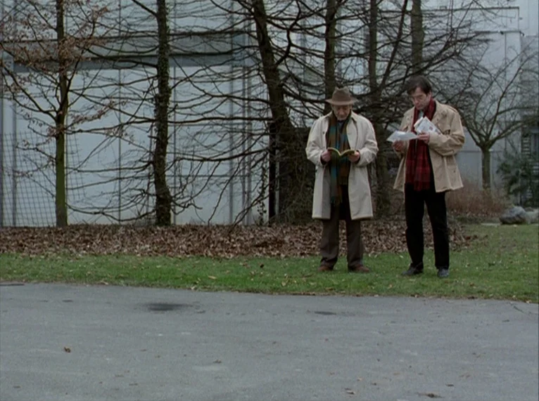 For Ever Mozart (1996) movie still: fullbody — Two men stand on a grass verge in front of bare, leafless trees. They are reading books and papers…