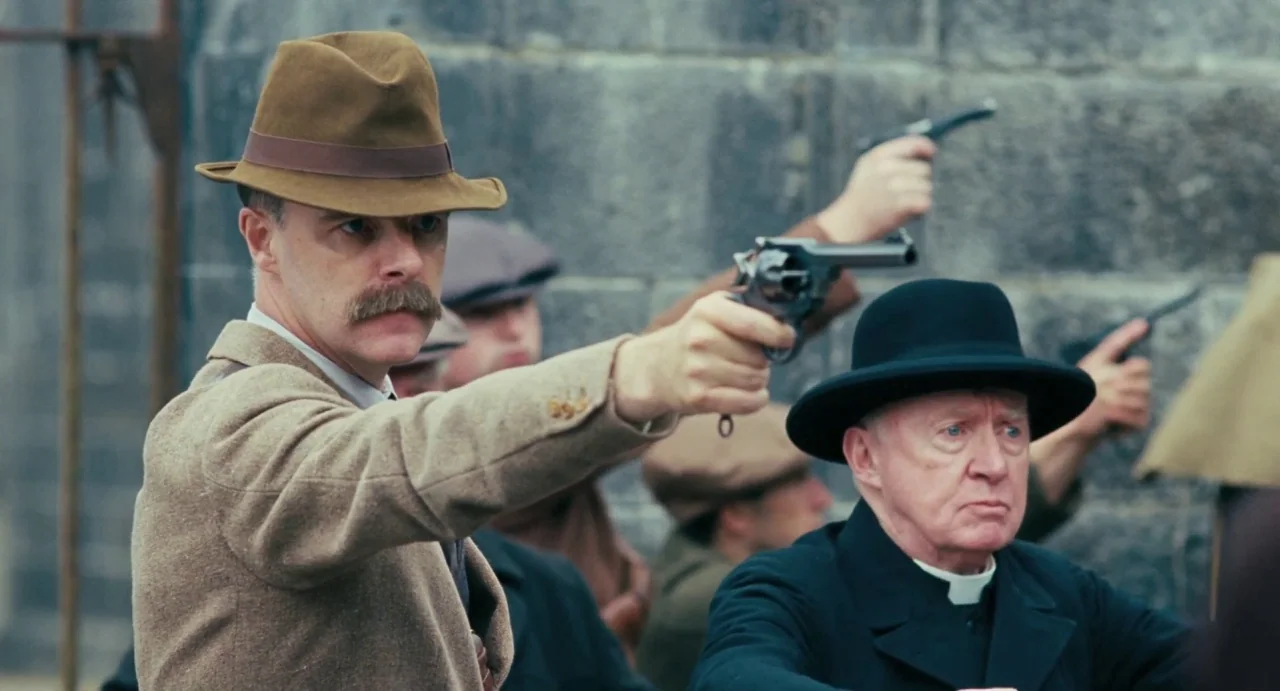 Jimmy’s Hall (2014) movie still: medium — A group of men stand before a grey stone wall holding firearms. A man in a tan hat and an elderly priest…