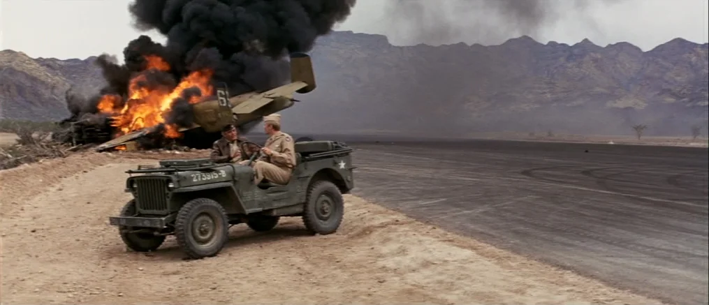 Catch 22 (1970) movie still: wide — A military airfield with a crash-landed burning airplane near a mountainous horizon. Two soldiers sit in a…