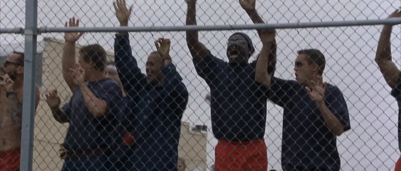 Bandits (2001) movie still: medium — A group of men stand outdoors behind a tall chain-link fence. The men raise their arms and cheer while wearing…