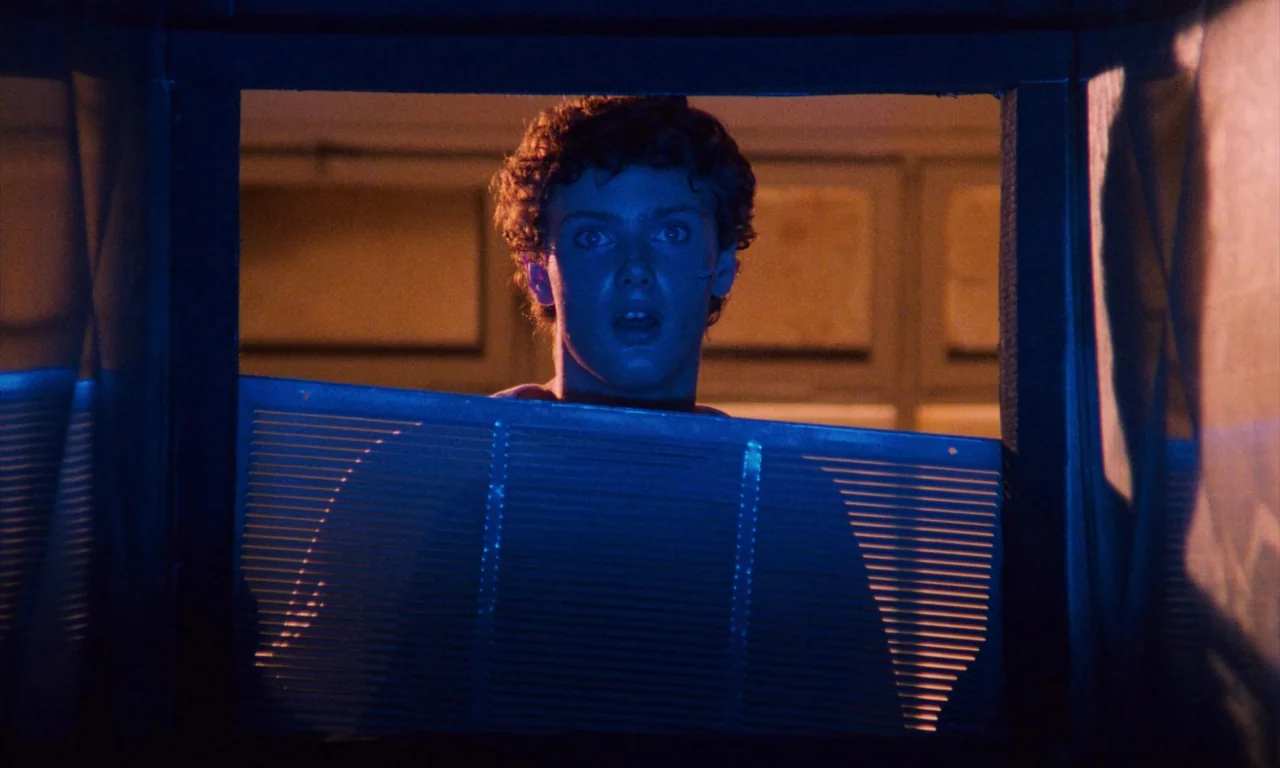 Demons (1985) movie still: closeup — A person looks through a framed window opening at night. Intense blue light illuminates their face against a…