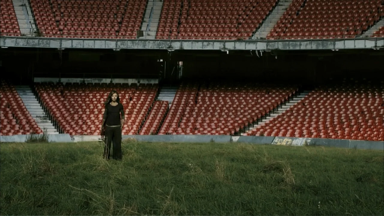 28 Weeks Later (2007) movie still: wide — A desolate stadium features rusted red seating rows overlooking a field of overgrown grass. A young woman…