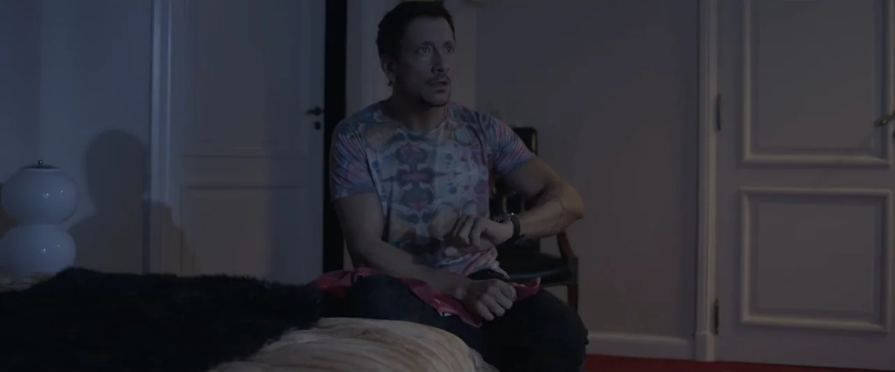 Perdida (2018) movie still: medium — A dimly lit room at night features a man sitting on a bed. He wears a patterned shirt and examines the time on…