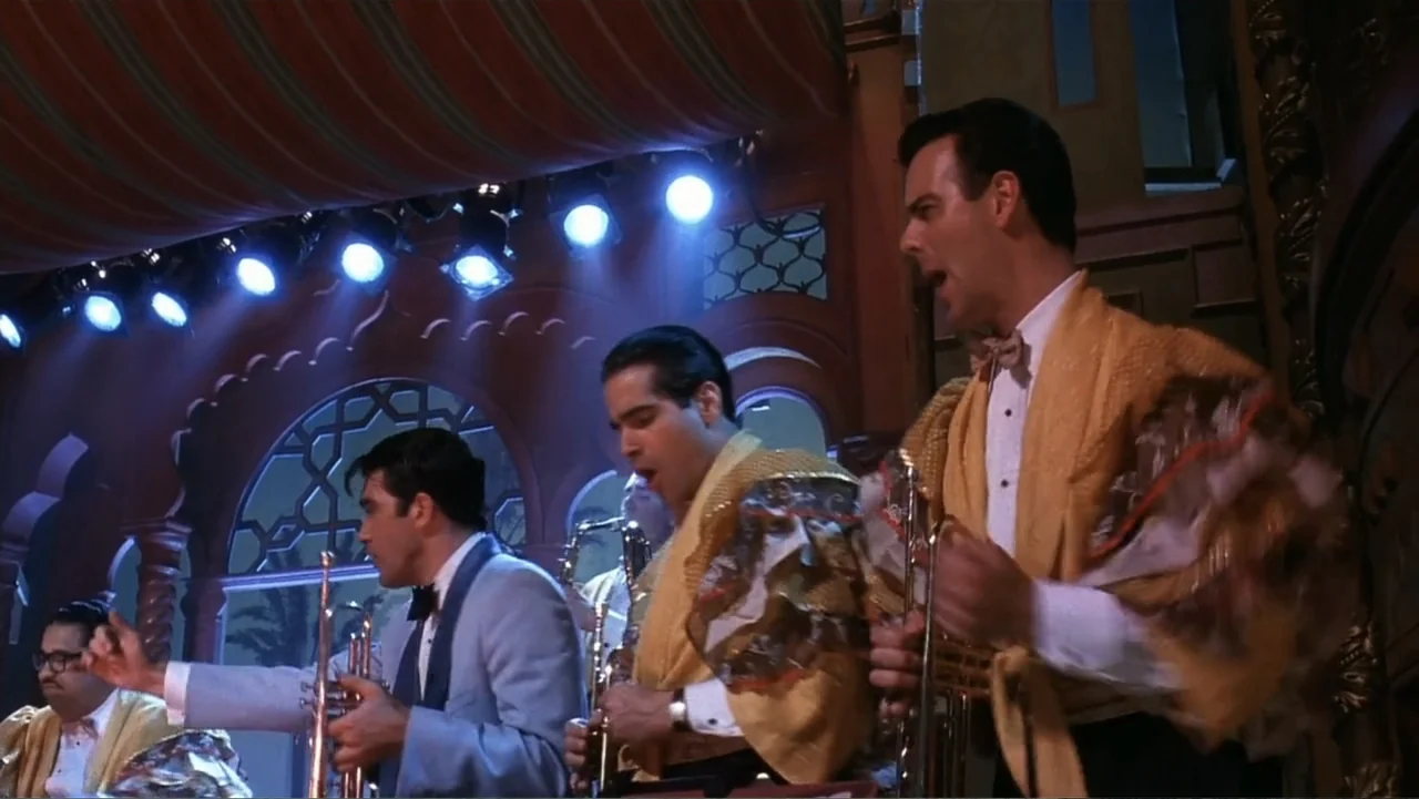 The Mambo Kings (1992) movie still: medium — A theatrical stage with ornate architectural arches and overhead drapery under bright spotlights. Three…