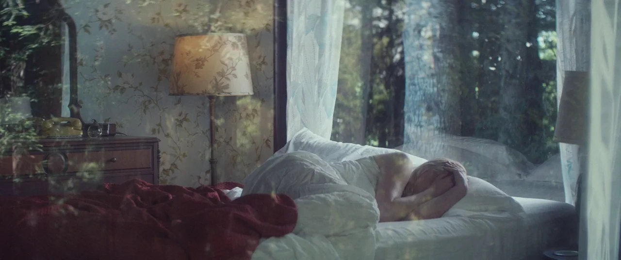 Woodshock (2017) movie still: medium — A bedroom features floral wallpaper and a wooden dresser with a yellow telephone. A person lies on a bed…