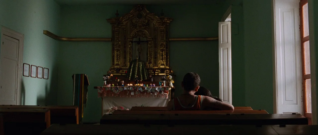 Sin Nombre (2009) movie still: wide — A quiet church interior features sage-green walls and a large, ornate gold altar piece. Two young people sit…