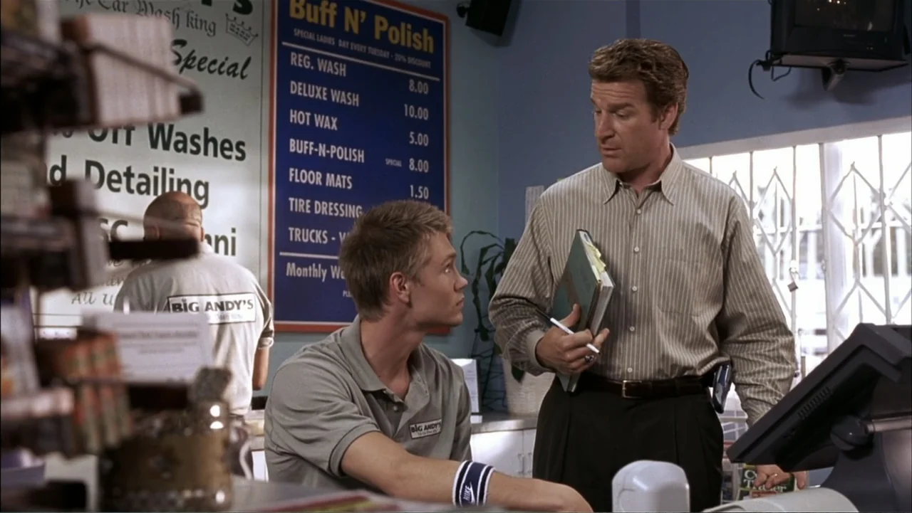 A Cinderella Story (2004) movie still: medium — A brightly lit car wash office with a blue pricing sign on the wall. A younger employee in a grey…
