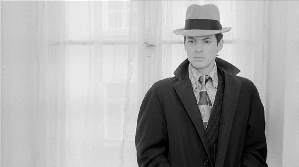 Love Is Colder Than Death (1969) movie still: medium — A man in a trench coat and fedora stands before a bright white window. The translucent…