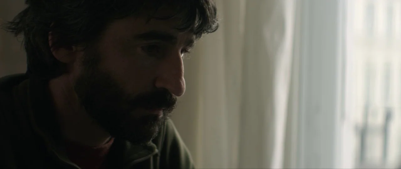 Silence (2012) movie still: closeup — A man stands near a bright window draped with translucent white curtains. He looks downward with a neutral…
