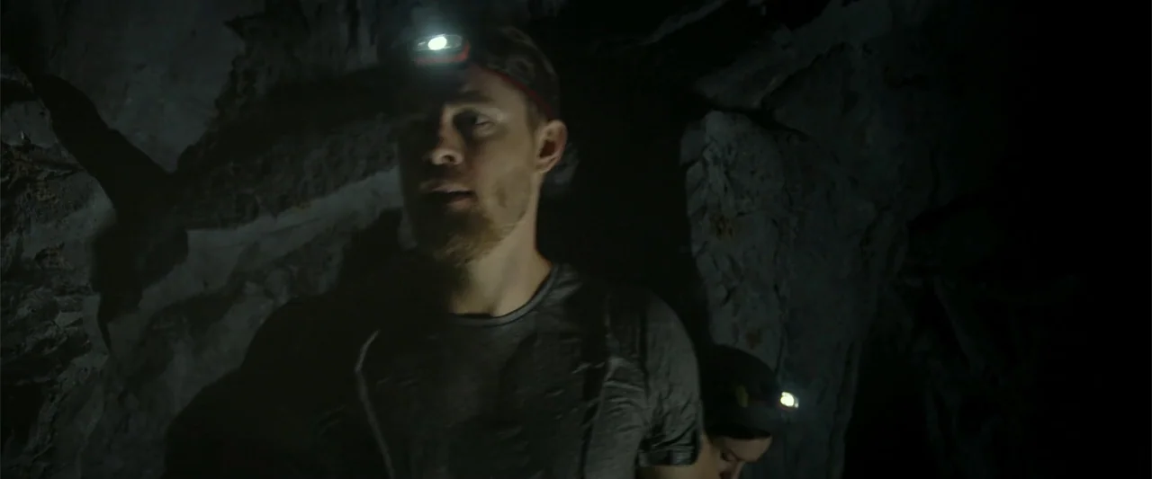 Black Water: Abyss (2020) movie still: medium — A dark rocky cave interior with textured walls illuminated by narrow beams of light. A man wearing a…