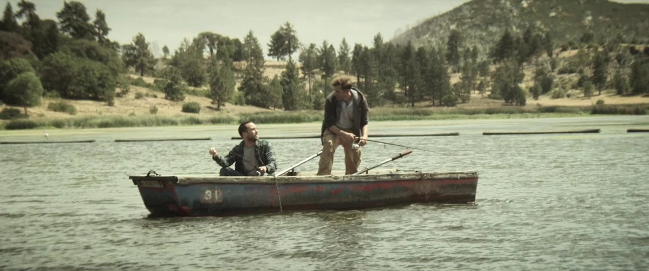 The Endless (2017) movie still: wide — A small weathered blue rowboat sits on the surface of a calm lake. Two men occupy the boat while holding…