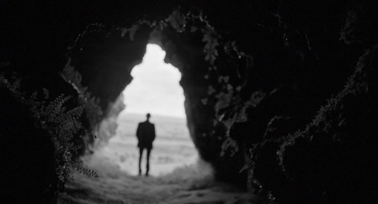 Song of Granite (2017) movie still: wide — A dark rocky sea cave opening frames a distant overcast sky and ocean. A lone man in a dark suit stands…
