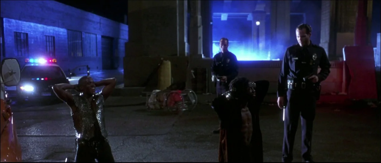 Strange Days (1995) movie still: wide — An industrial paved lot at night illuminated by harsh blue light and emergency vehicle strobes. Two men…