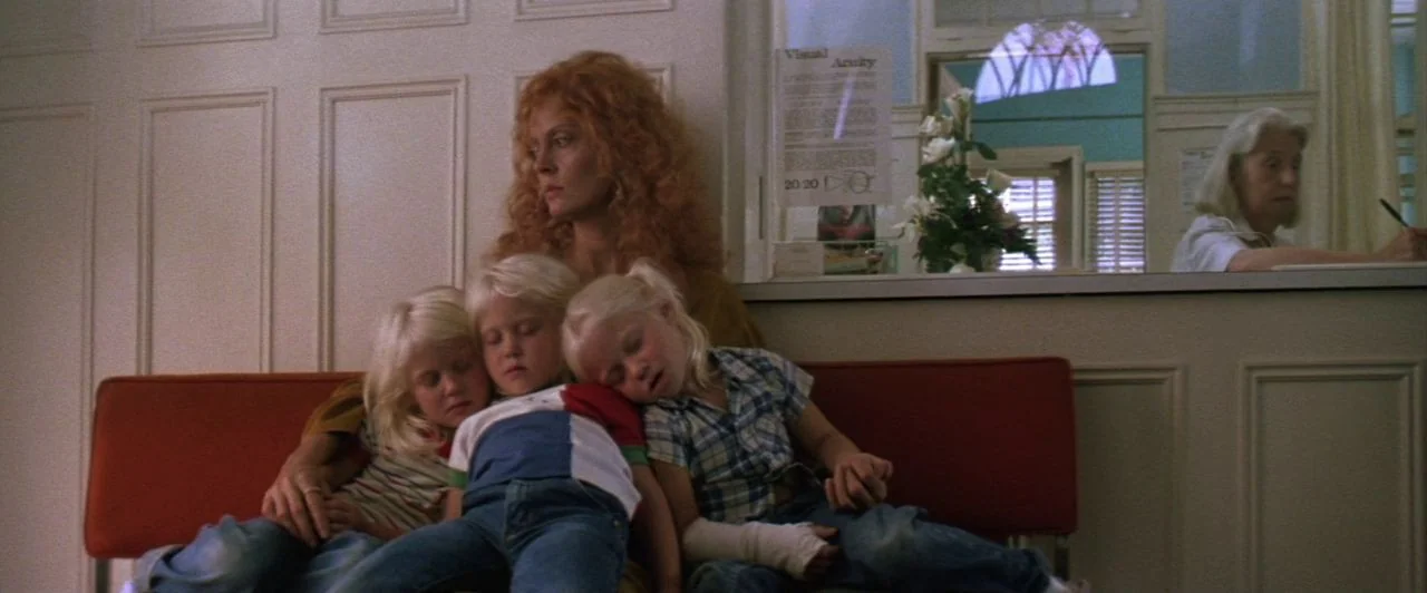 The Witches of Eastwick (1987) movie still: medium — A quiet medical reception area with beige panelled walls and a red vinyl bench. A woman sits…