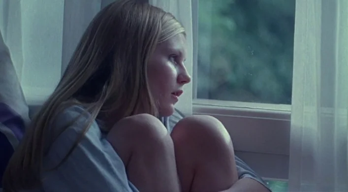 The Virgin Suicides (1999) movie still: closeup — A teenage girl sits against a window draped with thin white curtains. She has her knees pulled to…