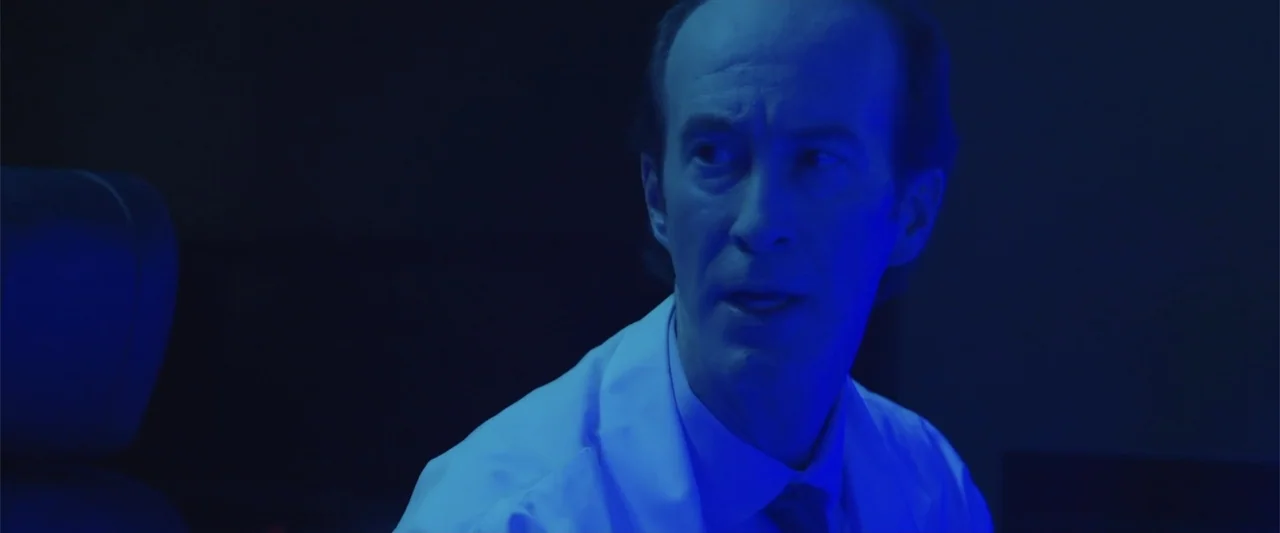 Presagios (2024) movie still: medium — A dark, sparsely lit laboratory space filled with shadows and blue light. A middle-aged man in a lab coat…