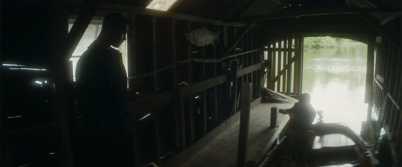 Black Bag (2025) movie still: medium — A rustic wooden boathouse with exposed framing overlooks a calm body of water. One man stands on the dock…