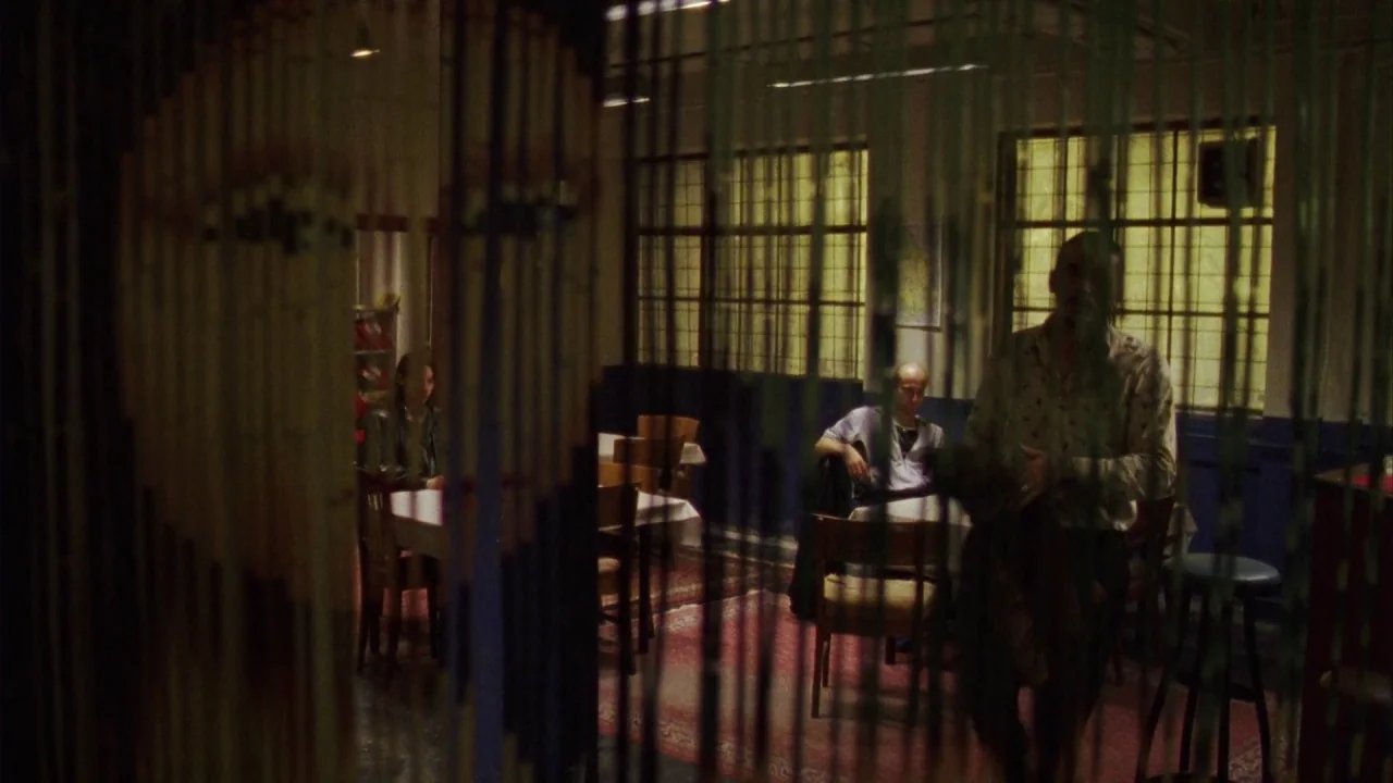 Pusher III (2004) movie still: wide — A dimly lit room with tables features green beaded curtains hanging in the foreground. Several men are seated…