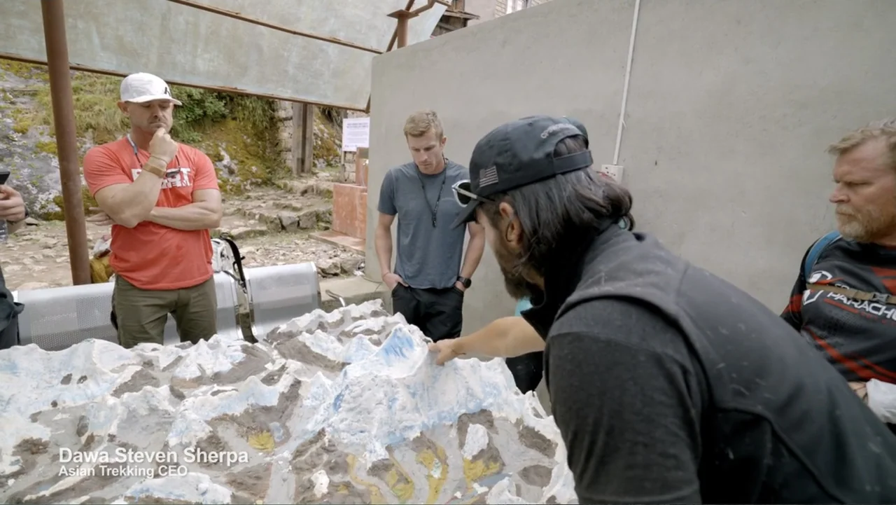 Drop Zone Everest (2023) movie still: medium — An outdoor concrete area contains a large white and blue scale topographical mountain model. Four men…
