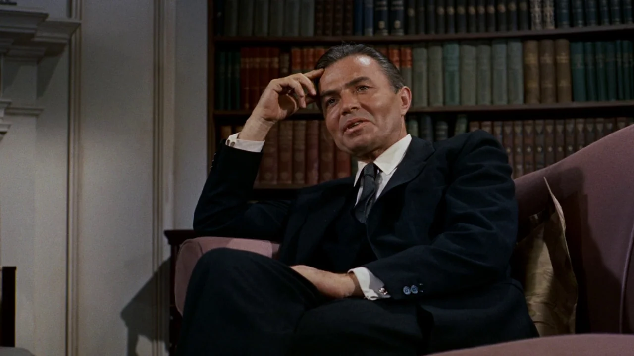 North By Northwest (1959) movie still: medium — A man sits in a room with a large wooden bookshelf in the background. He is wearing a dark suit and…