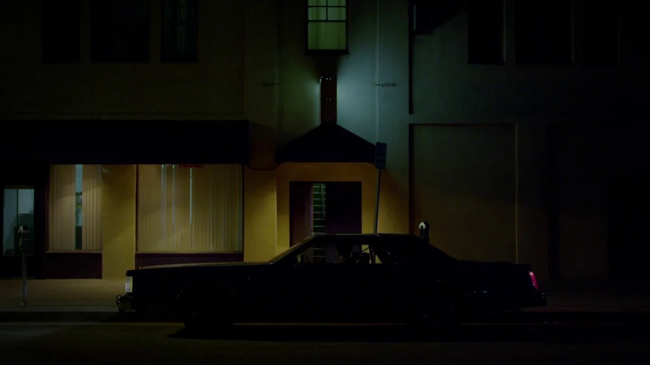 Suburban Gothic (2014) movie still: wide — A dark sedan sits parked at night in front of a commercial storefront building. A bright yellow light…