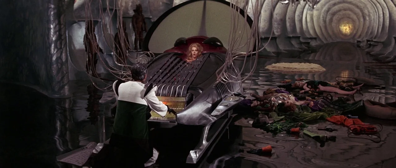 Barbarella (1968) movie still: wide — A dark subterranean chamber with curved walls and water covering the floor. A male figure operates a metallic…